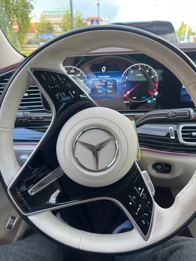 This media file shows a close-up view of a car's interior, specifically focusing on the steering wheel and dashboard. The steering wheel is covered in light-colored leather and features a prominent Mercedes-Benz logo at its center. Various controls are visible on the steering wheel, including buttons for cruise control (RES/SET+, SET-, CANCEL) and media control (OK, navigation arrows). The dashboard displays a digital instrument cluster with a speedometer, tachometer, and information about the car's status, including "После старта" (After start) and trip data like distance, fuel consumption, and time.
The central console has a gear selector with positions P, R, N, and D, and a start/stop engine button. The car's interior appears luxurious, with high-quality materials and ambient lighting. The setting is daytime, as indicated by the natural light coming through the windows, which also show a blurred view of trees and buildings outside, suggesting the car is parked or driving in an urban or suburban environment. The text "AIRBAG" and "MAYBACH" are visible on the steering wheel and trim, respectively, identifying the car as a Mercedes-Maybach. The overall impression is of a modern, high-end vehicle interior.
