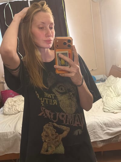 The media file is a selfie taken by a person in a dimly lit room. The subject is a young woman with blonde hair, looking directly at the camera. She is wearing a black t-shirt with a graphic print that appears to be a band or album cover, featuring a cat's face and a mythological creature. The text on the t-shirt reads "BE THE RATHER". She is holding a smartphone with an orange case, which is angled to capture the reflection. Her left hand is resting on her head, and she is wearing a delicate ring and a dark bracelet.
The background suggests a domestic setting, possibly a bedroom, with a bed covered in white bedding visible. The lighting is soft, and the overall mood is casual and intimate. The room appears to be in Vaganovo, Russia. The image is oriented sideways, suggesting it was taken with the camera held in portrait mode. There are no clear indicators of the time of day or weather conditions.
