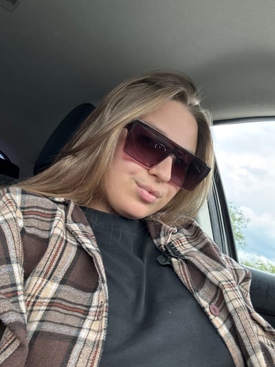 A person is sitting inside a vehicle. The person is wearing large, dark sunglasses, a black shirt, and a brown and tan plaid flannel. The location is not identifiable as Petrovka, Russia from this image. The background shows a car interior and a glimpse of the sky.