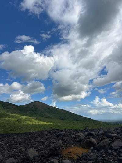 This image depicts a dramatic volcanic landscape in Rota, Nicaragua. The scene is divided diagonally, with the left side dominated by a vast expanse of a bright blue sky filled with fluffy white and grey clouds. The clouds are varied in texture and density, suggesting dynamic weather conditions.

The right side of the image transitions from the sky to a steep, green mountainside. The vegetation is dense and appears lush, indicating a fertile environment. This green slope is sharply contrasted by the foreground, which is covered in dark, rough volcanic rocks and scree. The rocks are of various sizes, and some patches of reddish-brown earth are visible amidst the black volcanic material.

The composition suggests a perspective from a high vantage point on the volcano, looking down and outwards. The lighting indicates it is likely daytime, with sunlight casting shadows and highlighting certain areas of the landscape, particularly on the green mountainside where a strip of bright sunlight falls. The overall mood is one of natural grandeur and rugged beauty. There is no visible text in the image.