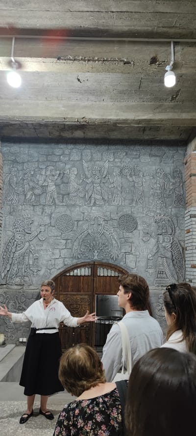 An indoor scene in Voskevaz, Armenia, features a tour guide gesturing with open arms towards a large, intricately carved stone relief on a wall. The relief depicts figures in historical attire, possibly ancient Armenian or Mesopotamian. The guide, a woman with short hair and wearing a white collared shirt and black skirt, speaks into a microphone. In front of her, a group of tourists, mostly seen from behind, listen attentively. The background includes a wooden double door and exposed concrete ceiling with lighting fixtures.
