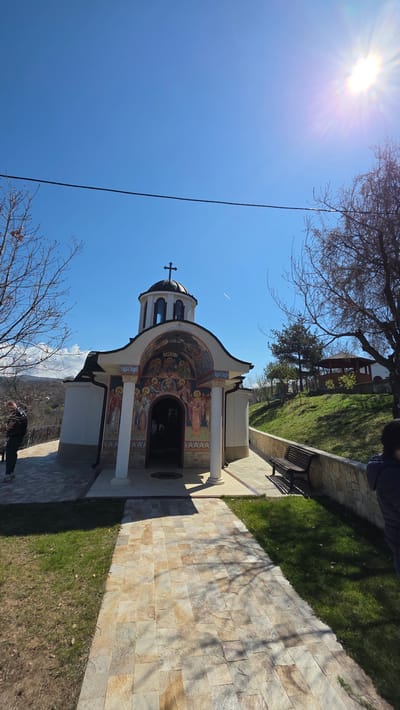 The image shows the St. Nicholas Church in Elenovo, Bulgaria, with the sun shining brightly from the upper left corner. The church is white with detailed painted murals around the entrance and a cross on top. A stone walkway leads to the church. To the right is a grassy area with a stone wall, and a wooden bench. There are trees in the foreground and background. A jet trail is visible in the sky. A person is visible in the bottom left and bottom right of the frame.
