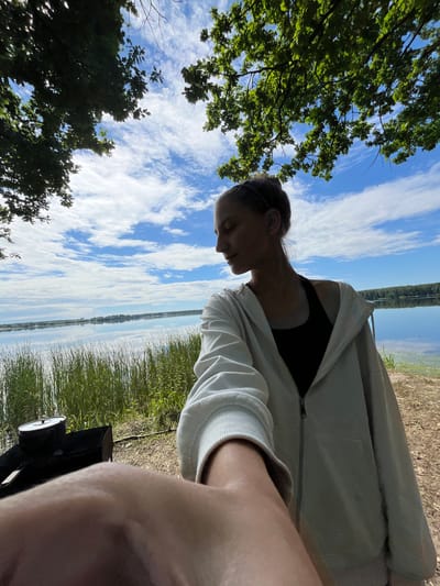 A young woman with her hair pulled back in a bun is captured in a selfie-style photo, looking down and to the left. She is wearing a light-colored hooded jacket zipped up over a dark tank top. The background shows a serene lake on a partly cloudy day, with tall green grass growing along the shore in the foreground. To the left, a portable grill with a pot on top is visible. The scene is outdoors, likely in a natural setting near a body of water, consistent with the rural landscape of Вяззеўскі сельскі Савет, Belarus, known for its lakes and greenery.