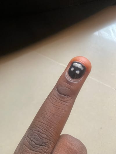 This image shows a close-up view of a person's index finger with a dark nail polish that has a few small, irregular white spots. The finger appears to be brown-skinned. The background consists of a light-colored surface with some shadowing. The lighting suggests it could be indoors or outdoors in daylight. There is no visible text in the image. The focus is on the detail of the nail polish. The specific activity or event is not clearly discernible, but it likely involves personal grooming or decoration. The location context provided is Wauro Leggal, Nigeria, which suggests the setting is likely a domestic or everyday environment within that region.