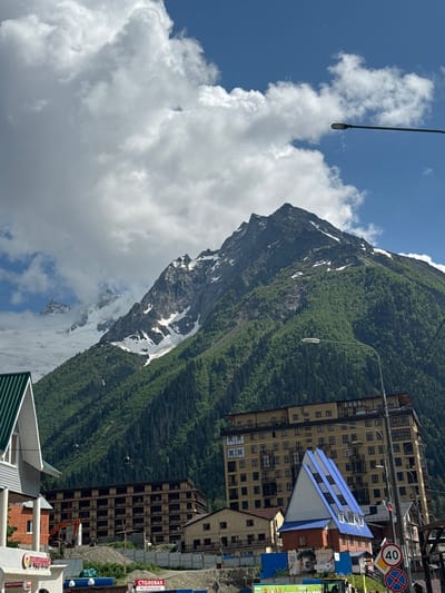 The image depicts a mountainous landscape with a village nestled at its base. Dominating the scene is a large, dark mountain face with patches of snow, indicative of a high altitude. The mountain is covered in lush green vegetation, suggesting a temperate season.

In the foreground, several buildings of varying architectural styles are visible. Some are modern, multi-story hotels with balconies and prominent blue roofs, while others appear older or under construction, with exposed concrete and an excavator nearby. A street sign with "40" in a yellow circle and a circular red sign with a diagonal bar are visible, suggesting a road and traffic regulations. Signs with Cyrillic text are also present, including one that reads "СТОЛОВАЯ" (dining room) and another with "ПРОДУКТЫ" (groceries).

The sky is a vibrant blue, with scattered white, fluffy clouds, indicating fair weather. The presence of a cable car system with cabins moving along cables suggests the area is a tourist destination, likely for skiing or mountaineering. A street lamp extends into the frame from the left, and a wire runs across the top of the image, possibly part of the cable car system or utility lines.

The overall impression is one of a scenic mountain resort town, capturing the juxtaposition of natural beauty and human infrastructure. The time of day appears to be daytime, given the bright sunlight casting shadows. There are no people clearly visible in the image.