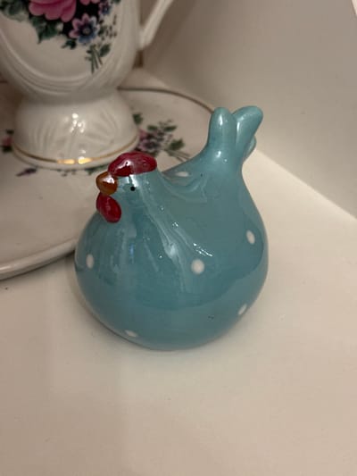 A static indoor scene in the city of Zelenogradskiy, Russia, displays multiple ceramic objects on a light-colored shelf. In the foreground, a glossy, teal-blue ceramic chicken figurine with white polka dots is positioned, facing left. The figurine features a prominent red comb and wattle, a small orange beak, and a black dot representing an eye. Its tail is raised and bifurcated. Behind and to the left of the chicken, a white ceramic teacup, or similar vessel, with fluted details and a gold-painted rim on its base, is partially visible. This vessel rests on a white ceramic saucer, also partially visible, adorned with a floral pattern consisting of pink, purple, and blue flowers with green foliage.