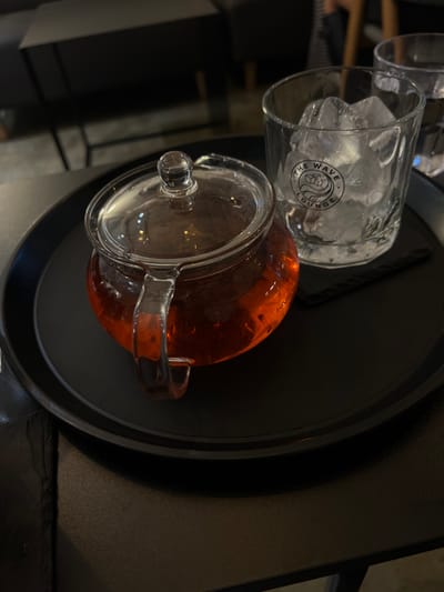 A black circular serving tray rests on a dark, textured table within a dimly lit indoor lounge, identified as The Wave Lounge, located in Mapo-gu, South Korea. On the left side of the tray is a clear glass teapot, filled with a reddish-brown liquid, consistent with brewed tea. The teapot features a transparent lid with a spherical finial and an integrated glass handle. To the right of the teapot, a clear glass tumbler is filled with irregular ice cubes. The tumbler displays a black printed logo that reads "THE WAVE LOUNGE" and incorporates a stylized cube icon. A thin, black stirring stick or straw is positioned horizontally on the tray between the teapot and the tumbler. The background is out of focus, revealing dark, indistinct furniture and a faint, partial silhouette of an individual, suggesting a public or commercial setting. No direct human interaction with the items is visible in the foreground.