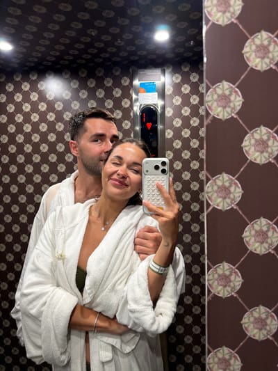 In the city of San Pellegrino Terme, Italy, two individuals, a man and a woman, are captured in a selfie taken inside an elevator. The woman is in the foreground, holding a smartphone and posing for the camera with a wink. She is wearing a white bathrobe over a green bikini top, adorned with necklaces and bracelets. The man is positioned behind her, with his head resting on her shoulder, also wearing a white bathrobe. The elevator interior features a patterned wallpaper with a geometric design and a touch panel for the elevator controls. The overall scene conveys a sense of intimacy and relaxation.