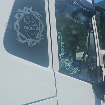 The image depicts the side of a white Volvo truck, showcasing decals with the Volvo logo on the windows. The upper window has a light blue Volvo logo with decorative swirls, while the lower window features a grey Volvo logo enclosed in a tribal-style circle. The location is assumed to be Le Perthus, France, based on the image description. The reflection inside the upper window shows a partial view of the interior and outside scenery.