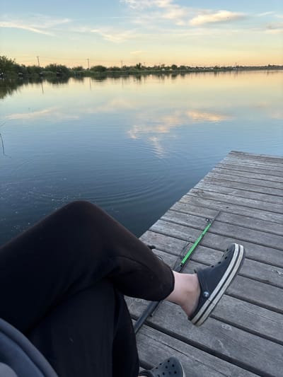 A person is sitting on a wooden dock, facing the water, likely fishing. The person is wearing black sweatpants and two Crocs sandals, one black with white stripes and the other gray. A green fishing rod is visible near the feet. The water is calm, reflecting the sky, which transitions from blue to a golden hue, indicating either sunrise or sunset. The opposite shore is lined with trees and possibly some buildings, reflecting in the water. The location appears to be a waterfront area in Khryashchevka, Russia.