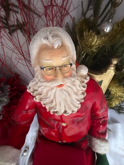 This image depicts a festive display located in Bagnoles de l'Orne Normandie, France. The central subject is a highly detailed, three-quarter view of a Santa Claus figurine, likely constructed from ceramic or resin, positioned slightly to the viewer's left. The Santa figure wears a red coat with sculpted white fur trim on the collar and cuffs, and small gold buttons on the front. He has a full, white, sculpted beard and mustache, rosy cheeks, and wears dark blue, rectangular-framed eyeglasses positioned on his nose. His gaze is directed downwards and to the left.
The background behind the Santa figurine consists of various Christmas decorations. To the left, thin, red, bare decorative branches are visible against a light background. To the right, a section of an artificial evergreen branch adorned with gold tinsel and a silver spherical ornament is present. A glittery gold object, possibly part of a sleigh, is partially visible behind Santa's right shoulder. A white, draped material, resembling artificial snow, forms the base around the figure, with another small silver ornament visible on it near Santa's left arm.