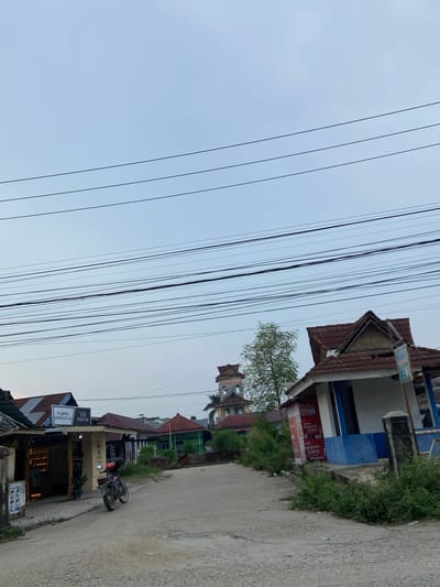 The image captures a street scene in Candirejo, Indonesia, characterized by a network of overhead power lines crisscrossing a pale, overcast sky. In the foreground, a dirt road winds through a collection of buildings with corrugated metal roofs. To the left, a shop labeled "WARUNG ENDOELITAH VN PERFUME" displays bottles of perfume. A motorcycle is parked on the side of the road near this shop. Further down the street, other structures of varying sizes and colors are visible, including a building with a blue and white facade displaying posters and an adjacent pillar. Lush green vegetation grows along the edges of the road and around some of the buildings. In the background, a multi-story building with a distinctive tower stands out against the horizon, hinting at the broader urban landscape of Candirejo. The overall impression is one of a typical, somewhat modest urban street in Indonesia, likely captured during the early morning or late afternoon given the soft light.