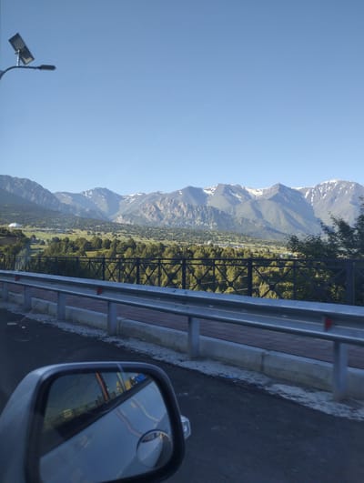 The image shows a roadside view with a mountainous landscape in the background. In the foreground, a car's side mirror reflects the road and surrounding scenery, including a metal guardrail and a pedestrian walkway. A solar-powered street lamp is visible on the left side of the image. The background features lush green hills dotted with trees, leading up to a range of snow-capped mountains under a clear blue sky. The overall scene suggests a scenic route outside a populated area. The image is not located in the city of Myk, Uzbekistan.