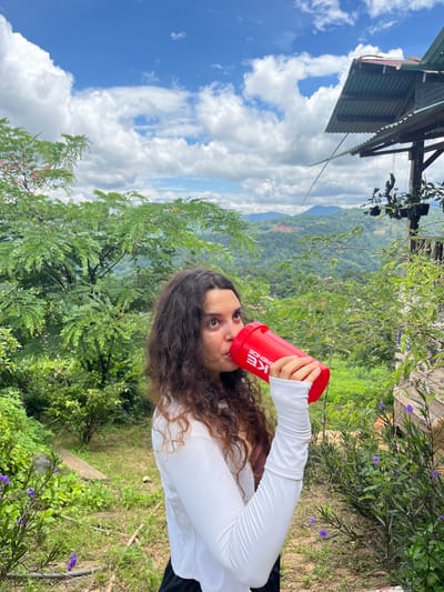A woman with long, dark, curly hair is shown from the chest up, drinking from a red shaker bottle. She is wearing a white long-sleeved shirt and black leggings. The background is a lush, green mountainous landscape under a blue sky with white clouds. A rustic wooden structure with a corrugated metal roof is visible to the right. The location is Sơn Điền, Vietnam.