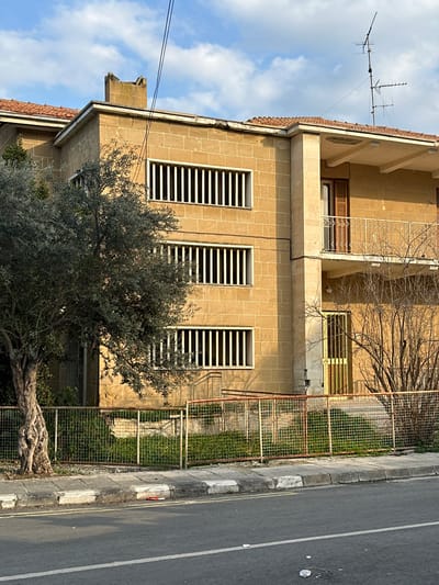 The image presents a two-story building in Agios Dometios, Cyprus, viewed from street level on a clear daytime. The building's exterior is constructed from yellowish-tan, rectangular stone-like blocks. The ground floor features three tall, narrow windows, each fitted with white frames and prominent horizontal louvers or blinds.

The upper floor includes a balcony with a simple, light-colored metal railing, behind which a wooden-shuttered window is visible. Another window, also with louvers, is partially visible on the upper floor to the right. An older-style television antenna is mounted on the building's tiled roof, silhouetted against a bright blue sky with scattered white clouds.

In the foreground, a large olive tree with a robust, gnarled trunk and silvery-green foliage occupies the lower left. To the right, a metal mesh fence delineates the property line, enclosing a patch of green ground and some bare-branched trees. Beyond the fence, a concrete sidewalk runs alongside an asphalt road. The road surface shows a white painted line and some light debris, including what appears to be a crumpled white cup.

The scene conveys a quiet, sunny day, likely late morning or early afternoon given the visible shadows. There are no people or discernible activities taking place. No specific text is visible in the image.