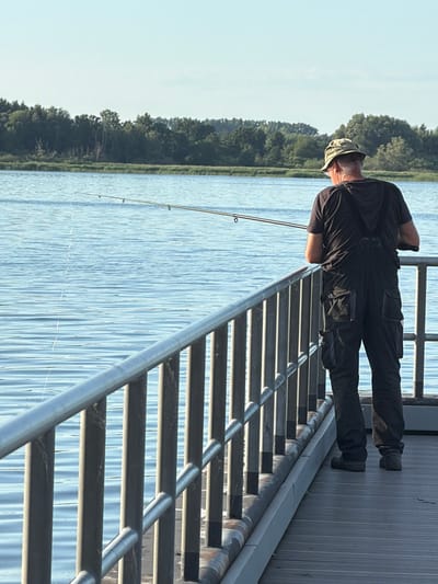An adult male is fishing from a pier on a body of water. He is wearing a black t-shirt, black overalls with multiple pockets, and a camouflage hat. He holds a fishing rod, with the line extending into the water. The pier has a metal railing and a wooden surface. In the background, there is a line of trees and a clear sky. The scene is set in Ribnitz-Damgarten, Germany.