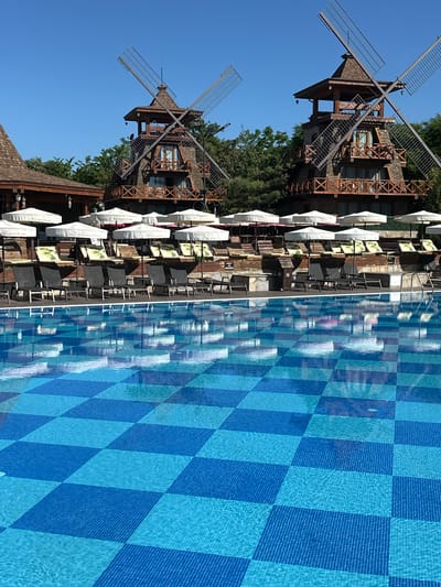 The image depicts a scene at Hotel Rila in Negushevo, Bulgaria. The left side showcases two windmill structures, likely part of the hotel's architectural design. Adjacent to the windmills, there is an outdoor seating area with chairs, tables, and white umbrellas. The right side shows a swimming pool, tiled with a blue and light blue checkered pattern. The sky above is clear and blue.