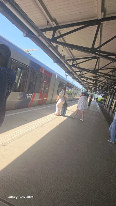The image depicts a train station platform in Vlaardingen, Netherlands, on a sunny day. A modern, silver and red train, identified by the "RET" logo, is stopped at the platform. Several people are visible. On the left, a person's back is to the camera, wearing a dark blue shirt and a black strap. Near the train, a person wearing a striped shirt and light-colored pants is standing next to a blue suitcase. Further down the platform, another person in a white shirt and shorts is walking away from the train, with their legs bent. More individuals are visible in the background, and a construction crane is seen in the distance against a clear blue sky. The platform is paved with a textured material, and a portion of a metal roof structure with exposed beams is overhead, casting shadows on the ground. The overall scene conveys a sense of travel and transit.