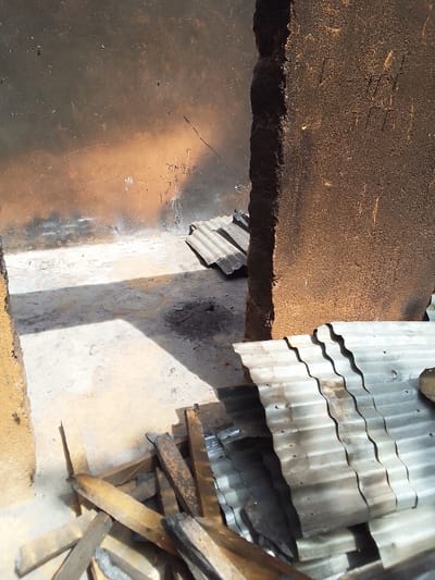 The image shows a scene of debris inside a building, likely a storage shed or outhouse, in the town of Kwal, Nigeria, located at coordinates 9.803 latitude and 8.646 longitude. The scene is captured from a low angle, showing the interior of the building. The walls are made of mud bricks and are blackened by fire. The floor is made of packed earth and covered in debris, including wooden planks and pieces of corrugated metal sheets. Some of the corrugated sheets lie flat on the floor, while others are stacked against a wall in the background. The light in the scene is coming from a window or opening in the wall. It is unclear what event occurred, but the debris suggests the building may have suffered some damage, possibly from a fire or storm. The scene is deserted with no people present.