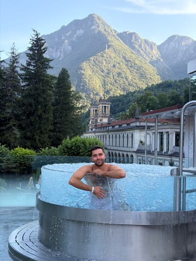 A man is partially submerged in a circular, bubbling hot tub with a transparent glass exterior, set against a backdrop of lush green mountains and a grand, classical building. The man, with dark hair and stubble, is looking towards the camera with a slight smile. He is wearing swim trunks. The hot tub's metal base features a ribbed texture, and water cascades down its sides. The building behind is ornate, with arched windows and a yellow tower. Tall evergreen trees are visible to the left of the building, and smaller trees and bushes form a line in front of it. The sky is a clear, pale blue. This scene is located in San Pellegrino Terme, Italy, likely at the Grand Hotel San Pellegrino or the associated spa.