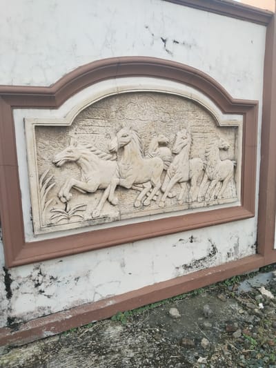 An outdoor scene in Bangsri, Indonesia, features a relief sculpture of horses embedded in a white wall. The sculpture, carved in a light, off-white material, depicts six horses in various dynamic poses, appearing to be running or galloping from left to right. The lead horse on the far left rears slightly, while the others follow closely behind, conveying a sense of movement and energy. Stylized plant motifs are visible on the left side of the relief, near the lead horse's front legs, and the background of the carving has a textured, rough appearance.
The relief is framed by a decorative border, which is light beige on the inside and a prominent reddish-brown on the outside. This framed section is set into a larger white wall, which shows significant signs of age and exposure to the elements, including numerous cracks, dark smudges, and patches of dirt or mold. The top of the wall is also bordered by a matching reddish-brown trim.
At the bottom of the image, the ground transitions from concrete to a patch of dirt and loose rocks, with some small green weeds growing sporadically. The lighting suggests it is daytime, with no shadows indicating a specific time, and the weather appears clear. There is no visible text in the image.