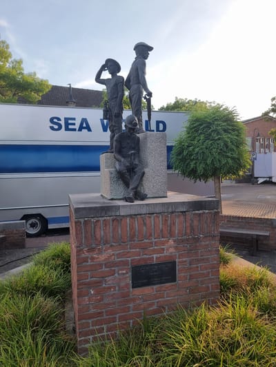 The image shows a statue of three miners in the city of Heusden-Zolder, Belgium. The statue is made of dark metal and depicts one miner sitting and holding a pickaxe, and two standing. One standing miner is saluting, and the other is holding a pickaxe. They are on a brick pedestal. Behind the statue, a large truck with "SEA WORLD" written on its side is parked. There is a small tree and a bench in the background, along with a brick building. The ground is paved with bricks. The sky is clear and blue, with some sunlight illuminating the scene. There is some greenery around the base of the statue.