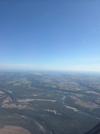 The image is an aerial view, taken from an airplane, of a vast landscape under a clear blue sky. The scene is dominated by a winding river that meanders through a densely forested area. On the left bank of the river, a small town or village is visible, characterized by a cluster of red-tiled roofs. Further in the distance, the landscape opens up to reveal agricultural fields and scattered settlements, with a hazy horizon suggesting a large urban area or the edge of a plain. There are no people or specific actions depicted in the image. The environment is natural with human settlements integrated into the landscape.