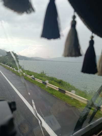 The image shows a view from inside a vehicle, looking out at a scenic landscape. In the foreground, blurred tassels hang from the vehicle's interior, obscuring part of the view. Through the windshield, a wet asphalt road with a white line runs alongside a body of water, likely a lake or reservoir. A metal guardrail with black and white markings separates the road from the grassy embankment. Lush green trees and rolling hills or mountains form the backdrop, fading into a hazy sky with clouds. The overall mood is serene and contemplative, suggesting a journey through a beautiful natural setting. The time of day appears to be overcast, possibly early morning or late afternoon. The location is identified as Tsamakaberd, Armenia. No text is visible in the image.