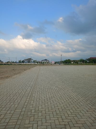 The image displays an expansive outdoor scene in Meureudu, Indonesia, under a partly cloudy sky. The foreground and midground feature a large, level surface paved with grey interlocking bricks arranged in a herringbone pattern, extending from the bottom of the frame towards the horizon. A faint vertical line traverses the center of this paved area. To the left of the paved section, an unpaved, sandy-brown dirt area is visible.

Along the distant horizon, a series of structures and natural elements are present. A prominent two-story building with a dark roof and light-colored walls is centrally located. To its right, a tall, white and red striped communication tower stands. Further right, a building with a reddish-brown sloped roof is partially visible. To the left of the central building, utility poles and power lines are discernible, along with a section of what appears to be a bridge or elevated roadway. Green trees and foliage are scattered along the horizon, both behind and between the structures. A large billboard with a red background and white text is visible near the central building. No individuals are present. A single dark-colored vehicle is faintly visible on a road in the distance, near the communication tower. The sky above is blue, covered by scattered white and grey cumulus clouds.