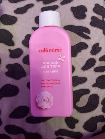 A pink bottle of "Café mimi" body lotion with a white cap is placed on a patterned fabric. The fabric has a spotted design, reminiscent of cow print, in black and grey on a light background. The lotion bottle features text in Russian, including "ЛОСЬОН ДЛЯ ТЕЛА ПИТАНИЕ" (Body Lotion Nourishing), and lists ingredients such as "ЭКСТРАКТ ПИОНА" (Peony Extract), "МАСЛО МИНДАЛЯ" (Almond Oil), and "ПАНТЕНОЛ" (Panthenol). A small pink floral design is visible on the bottle above the text, and a larger pink rose embellishment is present on the lower part of the bottle. The image is taken from a top-down perspective. The background pattern suggests the item is likely at a domestic setting within Shcheglovo, Russia.