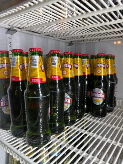 The image depicts a row of Ožujsko beer bottles arranged horizontally on a white wire rack, likely in a refrigerated display case. The bottles have yellow and red labels with the brand name visible. The location appears to be a retail environment, potentially in the city of Vitez, Bosnia and Herzegovina. The scene is static and there are no people visible in the frame. The primary objects are the beer bottles and the white wire rack.