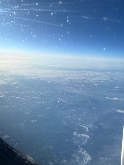 An aerial photograph depicts a vast, high-altitude landscape over Солгон, Russia. The upper two-thirds of the frame present a clear, bright blue sky with a subtle gradient to a lighter shade near the horizon, where faint contrails are discernible. Below the distinct but hazy horizon, a sprawling landmass is visible, characterized by irregular patches of white snow or low-lying clouds interspersed with darker ground, indicative of a winter environment. The bottom-left corner shows a portion of a dark aircraft window frame, possibly with frost or condensation. Numerous diffuse, bright, out-of-focus light artifacts or particles are scattered across the image, consistent with reflections or obstructions on the viewing window. No individuals or specific actions are depicted within the scene.
