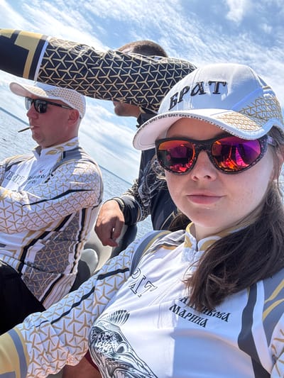 A medium shot captures three individuals on a boat, with two in the foreground and one in the background, on a sunny day with a cloudy sky. The person in the immediate foreground, a woman wearing a white baseball cap with black lettering that appears to say "БРАТ" and reflective sunglasses, is looking directly at the camera. She is wearing a white zippered jacket with a geometric pattern of gold and grey lines and star-like shapes. Next to her, to the left and slightly behind, is a man wearing a white baseball cap, sunglasses, and a similar white jacket with the same pattern. He is holding a cigarette in his mouth and looking to his left. In the background, a third person's arm is visible, wearing a black shirt with a gold geometric pattern. They appear to be leaning forward. The background shows a body of water, likely a river or lake, and a hazy sky. The location is identified as Городище, Russia, suggesting the boat is on a body of water near this city.