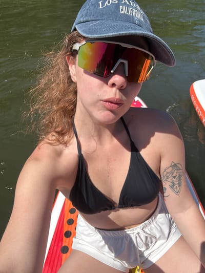 The image shows a young woman with reddish-brown curly hair wearing a black bikini top, white shorts, a denim baseball cap with "Los Angeles California" embroidered on it, and reflective sunglasses. She is sitting on an inflatable paddleboard, which is partially submerged in dark green water. The paddleboard has orange and white stripes with black circles. The woman is looking at the camera with her mouth slightly open. There is a tattoo of mountains and an airplane on her upper left arm. The scene is outdoors, likely on a river or lake, given the water and the inflatable paddleboard. The lighting suggests it is daytime and sunny. The location is identified as Ust-Mana, Russia, though the image itself does not contain any explicit visual cues to confirm this location.