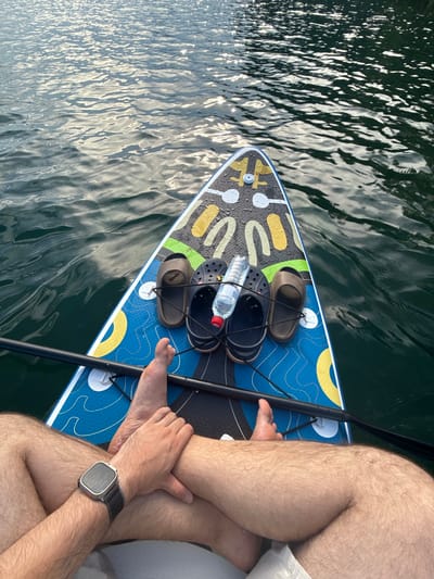 The image shows a person sitting on a paddleboard in the water. The person is wearing white shorts and a watch on their left wrist. Their legs are crossed. On the paddleboard, there are two pairs of sandals, a bottle of water, and a paddle. The water is dark and reflects the sky, indicating it is likely daytime. The scene appears to be a calm body of water, possibly a lake or calm sea, given the rippling surface. The overall impression is one of relaxation and outdoor activity. The watch on the person's wrist shows the time as 11:53. The location is Zaosinovo, Russia.