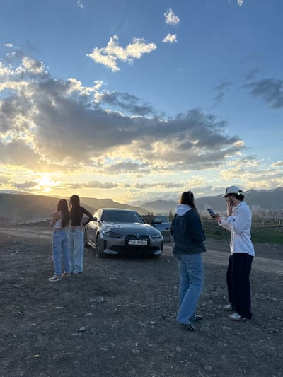 The image depicts a sunset scene with three people standing alongside a silver Citroen car on a dirt road. The location is on a hillside overlooking the city of Hrazdan, Armenia. The sky is filled with clouds and the setting sun.
The person furthest from the camera is an adult male wearing a white cap, a white button-down shirt, and dark trousers; he is holding a cell phone and looking at it. Closer to the camera is an adult female wearing a denim jacket and blue jeans. Two younger females are standing near the front of the car, both wearing tops and jeans. The car has a license plate, "66 MH 066." The background consists of a mountain range.
