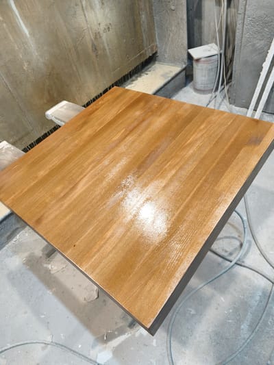 The image displays a square wooden tabletop undergoing a finishing process within an industrial workshop in Negorci, North Macedonia. The tabletop, positioned centrally, exhibits a reddish-brown, medium-toned wood grain, coated with a wet, glossy sealant that reflects a bright light source on its surface. It rests on two light-colored, rectangular support blocks positioned at its front and rear edges. The surrounding environment consists of unfinished grey concrete walls, one with vertical streaks and a dark base line, and another featuring a recessed section. The concrete floor is mottled with various light and dark patches, covered by coiled grey cables or hoses. In the background, a white metal barrel with faint red banding is visible, topped by a white rectangular lid, situated against a concrete structure. No individuals are present in the scene.