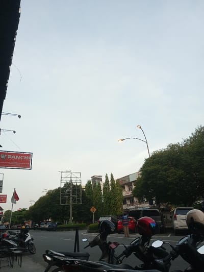 An urban streetscape in Pontianak, Indonesia, is depicted under an overcast, light grey sky.
In the immediate foreground, a dark architectural overhang partially obscures the upper left corner. Below it, a rectangular red sign displays "FANCHI INDONESIA" with a stylized bird logo. At the bottom, several scooters are parked facing right, with visible helmets in red, black, and beige. "Honda" branding is present on some vehicles. Partially visible black metal chairs or stools are on the far left.
The midground features a paved road or parking area with a curved concrete median. On the right, a red hatchback car is parked, with a male individual in a blue shirt and light trousers standing beside its open rear hatch. A dark SUV and a white SUV are parked further behind. A tall, dual-arm streetlamp with illuminated orange-yellow bulbs extends from the right. A large, empty metal billboard frame stands centrally on the median, near a yellow diamond-shaped road sign. An Indonesian flag (red and white) flies on a pole to the left of the median.
The background includes a multi-story building with a light-colored, weathered facade and multiple windows on the right. Numerous trees, including large canopy trees and slender conical evergreens, line the street and the median, providing dense green foliage throughout the scene.