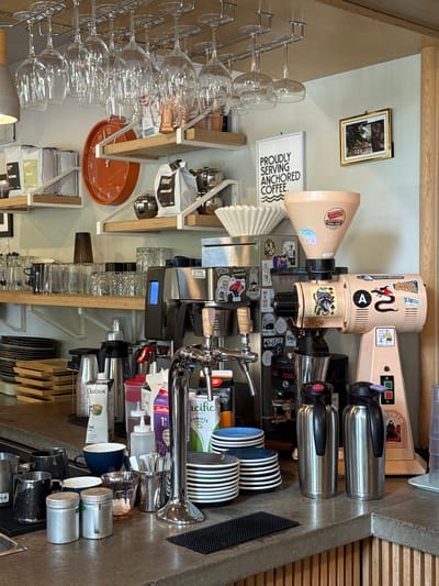 This indoor shot shows a countertop and the items placed on and near it. A coffee grinding machine with stickers is the central object. Next to it are two stainless steel containers. Several stacks of cups are next to the containers. A chrome coffee tap stands among them. Further to the left are various items such as milk cartons, coffee syrups, and more stacks of cups. Shelves with various items are stacked higher up on the wall. A wine glass rack holding several glasses is mounted on the wall above the shelves. A clock, coffee packets, and a picture are also seen on the shelves. Signs that read "PROUDLY SERVING ANCHORED COFFEE" and a frame is mounted on the wall. The location or the city of Chelsea, Canada is unknown from the image. No people are seen in the image.