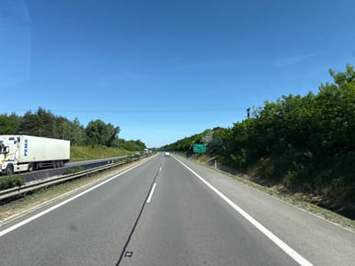 The image depicts a highway in Lozorno, Slovakia, on a clear, sunny day. The view is from inside a vehicle, likely a car or truck, traveling on the right-hand lane of a multi-lane road. A white PEMA semi-trailer truck is in the adjacent lane to the left. The road is asphalt with painted white lines marking the lanes. A sign on the right side of the road indicates distances to Brno (100km) and Malacky (12km). The landscape on either side of the road consists of trees and vegetation. In the distance, other vehicles are visible on the highway.