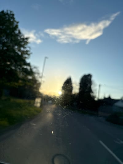 The image shows a view through a dirty car window. Outside, a blue sky with a few clouds is visible. Trees and other vegetation are blurred in the background. The sun is visible and appears to be setting. The location is Oswestry, United Kingdom. A road and a streetlamp can also be seen.