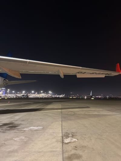 The image displays an airport tarmac at night, captured from a unique perspective alongside an aircraft wing. The dominant feature is the large, light-grey wing of an airplane, stretching across the left side of the frame with a striking red wingtip. Prominently displayed on the wing's surface is the registration number "RA-54918," indicating a Russian-registered aircraft.

To the right, a vast concrete apron extends into the distance, illuminated by numerous bright lights that highlight a long line of parked aircraft. The dark night sky hangs above, suggesting clear weather conditions. The concrete ground below the planes exhibits various stains and marks from operations. A small red and white traffic cone is visible on the apron in the lower right. The scene depicts a quiet yet operational atmosphere at a major airport after dark, with no specific activity or people discernible. Given the location context of Sdot Dan Regional Council, Israel, this scene likely takes place at Ben Gurion Airport (TLV).