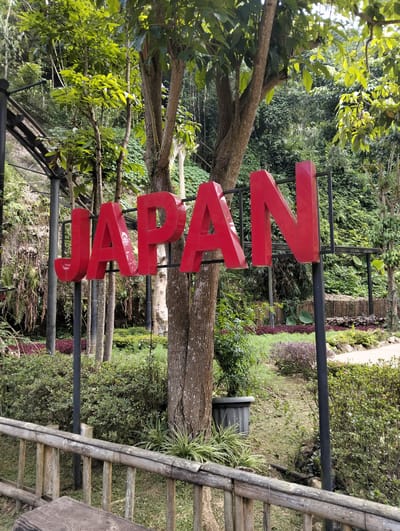The image shows a bright red sign spelling out "JAPAN" in large, three-dimensional letters. The sign is erected outdoors, in what appears to be a park or garden setting, surrounded by lush green foliage and trees. The lettering is bold and casts shadows, indicating the presence of sunlight. In the foreground, a rustic bamboo fence runs horizontally across the bottom of the frame. Behind the sign, a dense wall of green vegetation, including trees and various plants, forms the backdrop. Some of these plants appear to be in raised beds or planted in a landscaped manner, with some areas of reddish-purple foliage visible. The overall scene is one of an outdoor attraction, possibly a themed area within a larger park, located in Gudangkahuripan, Indonesia.