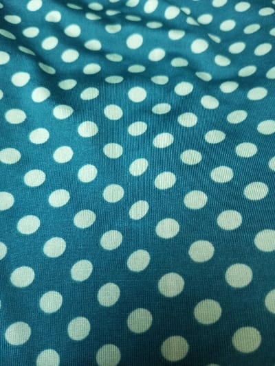 The image displays a close-up of a blue fabric with a pattern of uniformly spaced white polka dots. The texture of the fabric appears to have a subtle ribbing effect. The lighting is somewhat uneven, causing slight variations in color across the fabric surface. The polka dots maintain a consistent size and shape throughout the frame.