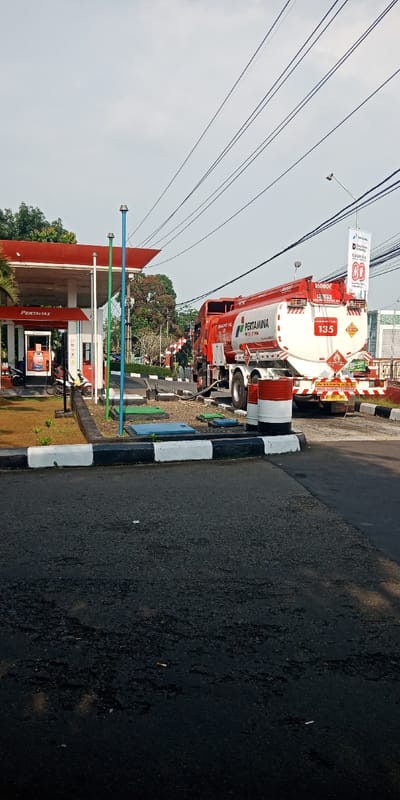 The image depicts a fuel station, specifically a Pertamax-branded station under the Pertamina company, located in Cijedil, Indonesia. A red and white Pertamina fuel tanker truck, marked with "16000L" capacity and a hazard sign "135," is parked adjacent to the station's fuel storage area. A hose extends from the tanker to an underground fuel tank access point, indicating an ongoing fuel delivery operation.
The gas station features a red-roofed canopy covering multiple fuel pumps, at least two of which are visible. One orange-colored fuel dispenser is clearly visible. Two motorcycles are parked near the pumps, with at least one person visible near a motorcycle, possibly a customer or attendant.
The immediate foreground shows an asphalt pavement with a black and white striped curb. Beyond the curb, a raised concrete border encloses an area with multiple square and rectangular access panels covering underground tanks, alongside three tall, slender ventilation pipes (one white, one green, one blue). A small patch of short, sparse grass with young plants is present.
Overhead, numerous power lines traverse the sky, which is a light, overcast gray. A street light pole with a white banner displaying Pertamina branding is visible on the right side of the frame. In the background, green trees and other structures are visible, including what appears to be a red and white striped barrier, possibly at an entrance or exit point. The overall scene suggests a routine daytime operation at a fuel station.