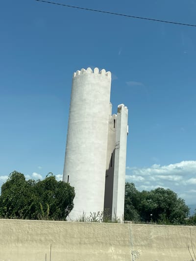 The image shows a white, crenellated tower that appears to be made of concrete or stucco. The tower is cylindrical and leans slightly to the right. To the right of the main tower, there is a narrower, rectangular structure attached to it, also with crenellations at the top. The tower is set against a clear blue sky with scattered white clouds. In the foreground, there is a rough, textured concrete wall. Behind the wall and to the left of the tower, there are lush green trees and foliage. The image appears to be taken from a slightly elevated position, possibly from a moving vehicle, as indicated by a faint horizontal line across the top of the image, which could be a reflection or a power line. There are no people visible in the image. The environment suggests an outdoor, possibly rural or semi-urban setting, given the presence of the tower and the natural vegetation. The location is identified as Berbuki, Georgia.