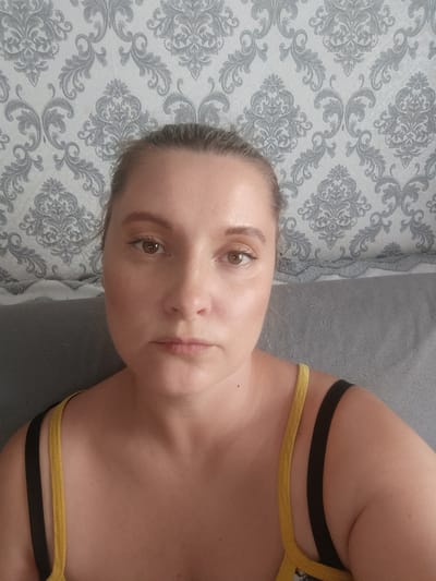 A woman is captured in a medium close-up, selfie-style portrait indoors. She has light-colored hair pulled back, brown eyes, and a neutral to slightly serious expression as she looks directly at the camera. Her skin appears fair, and she is wearing a yellow top with a black strap visible over her left shoulder.
Behind her, a wall is covered with a grey and white patterned wallpaper, featuring an ornate, damask-like design. Below the wallpaper, a section of a grey fabric sofa or cushioned headboard is visible, extending across the lower portion of the background. The lighting is bright and even, suggesting an indoor setting during the daytime or a well-lit interior. No specific activities are taking place beyond the subject posing for the photograph. There are no visible location cues within the image itself. No text is visible in the media. Based on external information, the setting is a Closed administrative–territorial formation "Mezhgorye", Russia.
