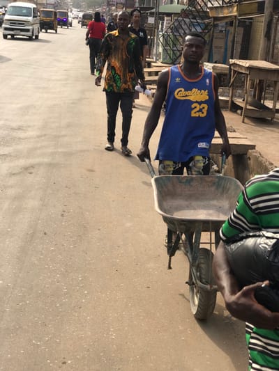 This image depicts a street scene in Umuokpu, Nigeria, under bright daylight. In the immediate foreground, partially visible on the right, is a person wearing a green and white horizontal striped shirt, holding a black, wrapped object. The central figure is a dark-skinned male, facing forward, wearing a blue Adidas "Cavaliers 23" basketball jersey and black shorts with yellow cartoon character graphics. He is pushing an empty, well-used metal wheelbarrow on the paved road surface. Directly behind him, another dark-skinned male walks, wearing a long-sleeved shirt with a green, orange, and black abstract pattern, dark trousers, and sandals, holding a rolled-up white paper in his left hand. Further down the street, several other individuals are walking, including a person in a red shirt. The road is paved with a light-colored material, showing some dirt and wear. To the right, an elevated, uneven sidewalk or ground area with a narrow drainage channel separates the road from market stalls or informal structures made of wood and metal. In the background, a line of buildings, some with corrugated roofs and visible signage, borders the street. Multiple vehicles, including a white van and yellow auto-rickshaws (kekes), are present on the road in the distance, indicating traffic.