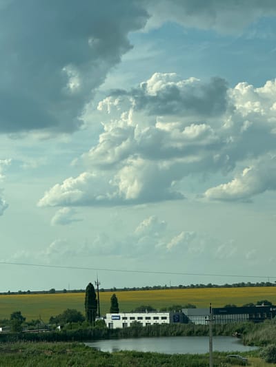 The image depicts a broad, natural landscape under a dynamic sky. Dominating the upper portion of the frame are large, dramatic cumulus clouds, varying in color from bright white to deep gray, suggesting a mix of sunlight and shadow. The lower portion of the image shows a body of water, possibly a lake or pond, with lush green vegetation along its banks. Beyond the water, a flat, rural landscape unfolds, characterized by a vast field of yellow flowers, likely sunflowers, stretching towards the horizon. A few trees and a building complex are visible on the right side of the frame, near the water's edge. The building appears to be industrial or commercial, with a sign that reads "БЕЛТРАНС" and another partially visible logo. Power lines traverse the scene horizontally, dividing the sky from the land. The overall impression is one of a rural, possibly agricultural, setting under a dramatic, cloud-filled sky. The image does not provide specific details that would definitively place it in Bartolomeevskii, Russia.