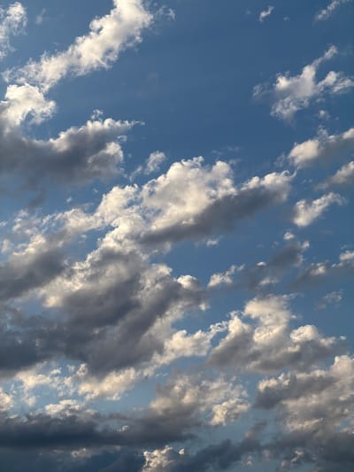 The image shows a sky filled with a mix of white and gray clouds against a bright blue sky. Some clouds are wispy and translucent, catching the sunlight, while others are darker and more substantial, suggesting depth. The overall impression is of a dynamic sky with varying cloud formations. The image does not depict any urban elements, people, or specific activities, and therefore cannot be localized to Arcahueja, Spain, or any other city.